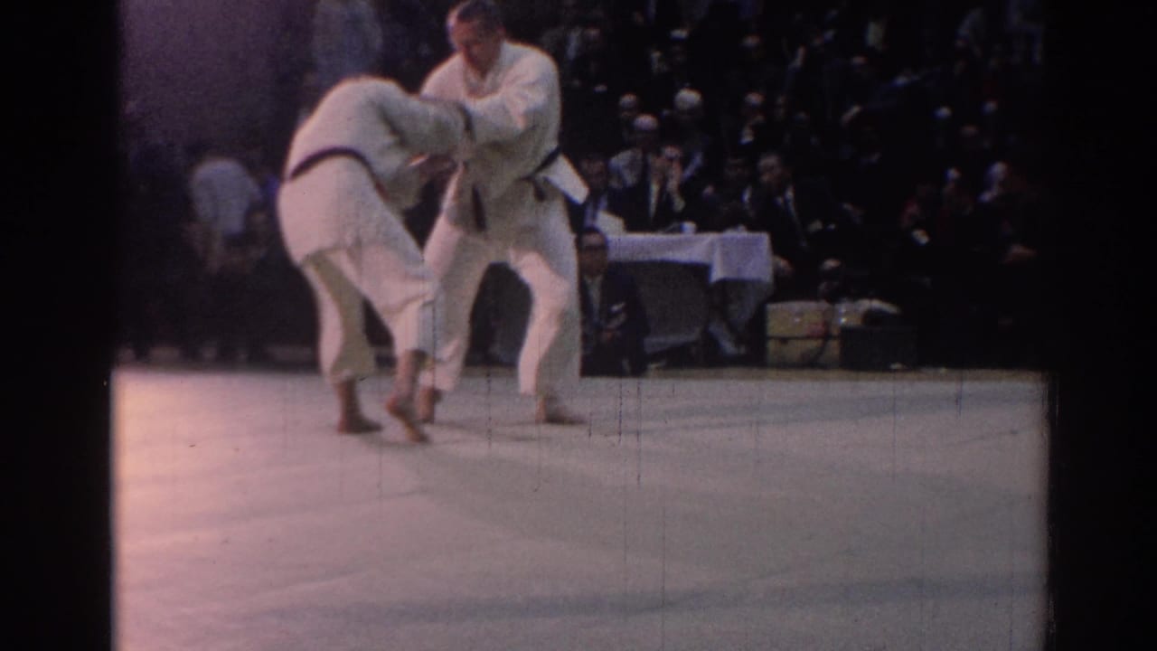 Two white-gi karate competitors face each other on the mat in Los Angeles, with spectators and a table official in the background.