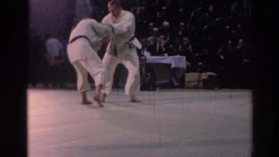 Two white-gi karate competitors face each other on the mat in Los Angeles, with spectators and a table official in the background.