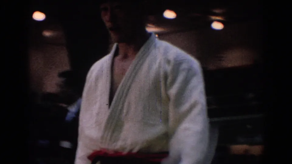 1967 Los Angeles 8mm frame showing a judo competitor in a white gi and red belt near the edge of an indoor match area.