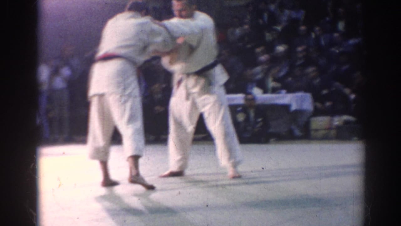 Two judo athletes in white gi engage during a 1967 Los Angeles indoor match, with spectators visible beyond the mat.
