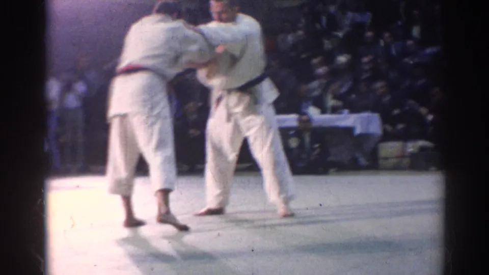 Two judo athletes in white gi engage during a 1967 Los Angeles indoor match, with spectators visible beyond the mat.