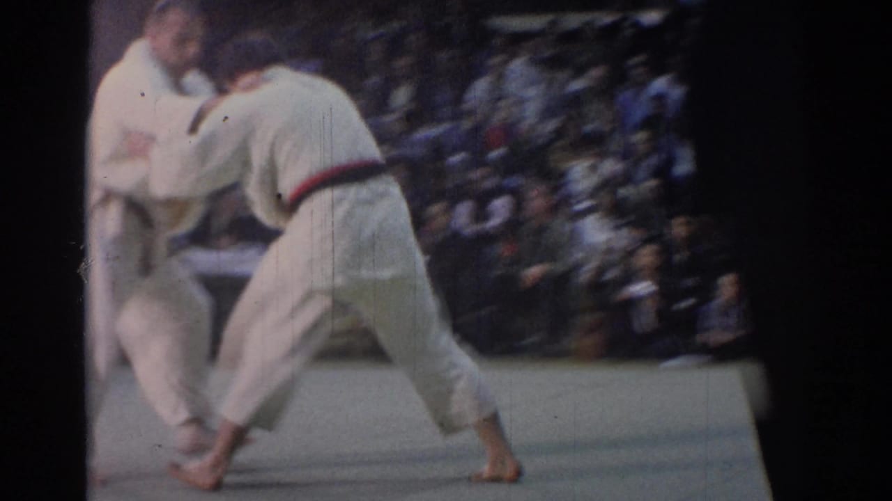 A 1967 Los Angeles judo match continues on a pale mat, with two competitors in white gi moving before a seated audience.