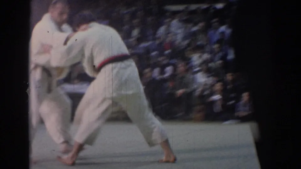 A 1967 Los Angeles judo match continues on a pale mat, with two competitors in white gi moving before a seated audience.