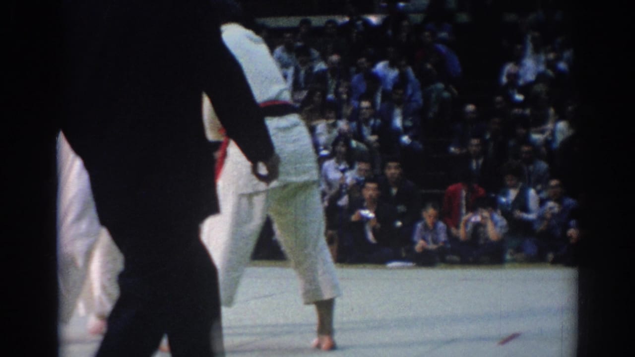 Indoor 1967 Los Angeles judo scene with an official in dark clothing, a competitor in a white gi, and seated spectators in the background.