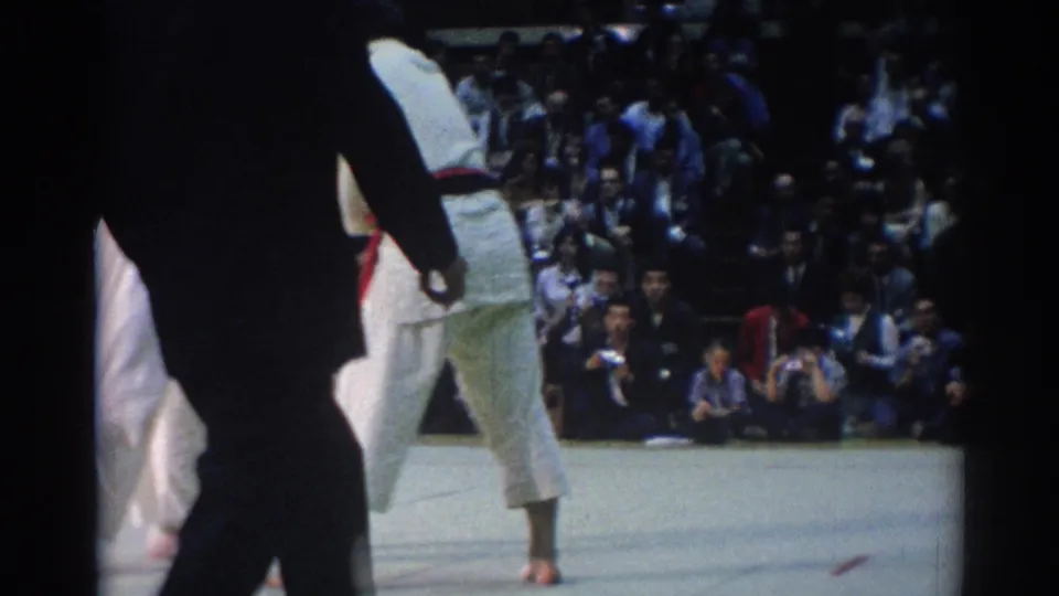 Indoor 1967 Los Angeles judo scene with an official in dark clothing, a competitor in a white gi, and seated spectators in the background.