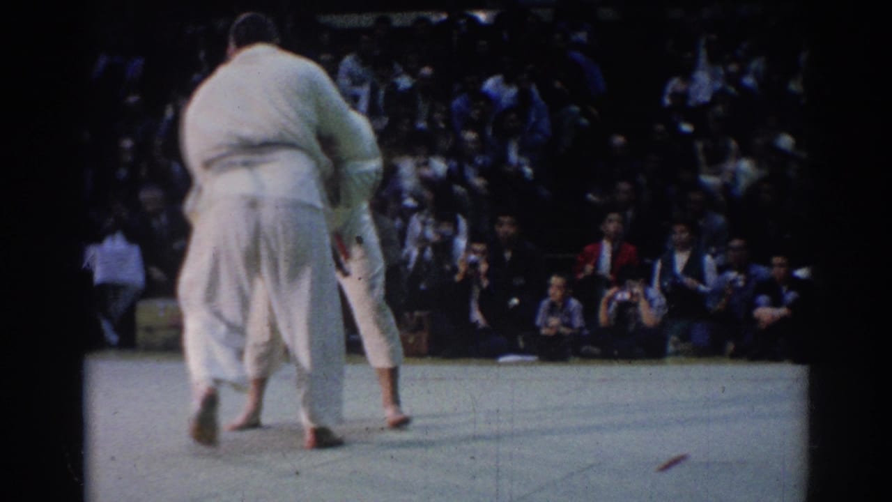 Two barefoot judo competitors grapple on a mat before a seated audience.