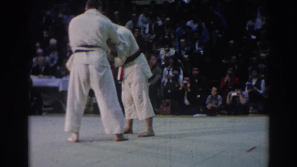 Two men in white judo uniforms hold a standing grip during the 1967 Los Angeles match, with spectators and cameras behind them.