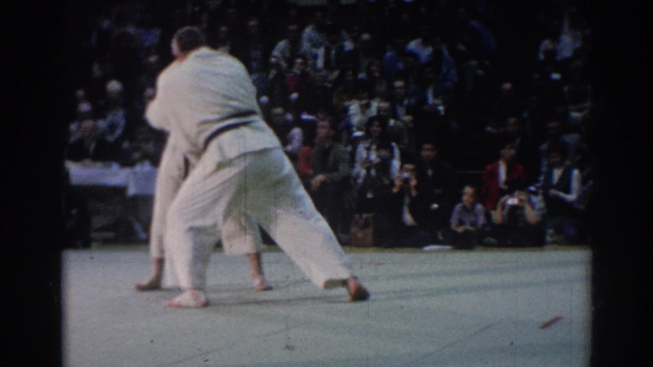A judo exchange unfolds on the indoor mat, with one competitor leaning into the other before a seated crowd in Los Angeles footage from 1967.