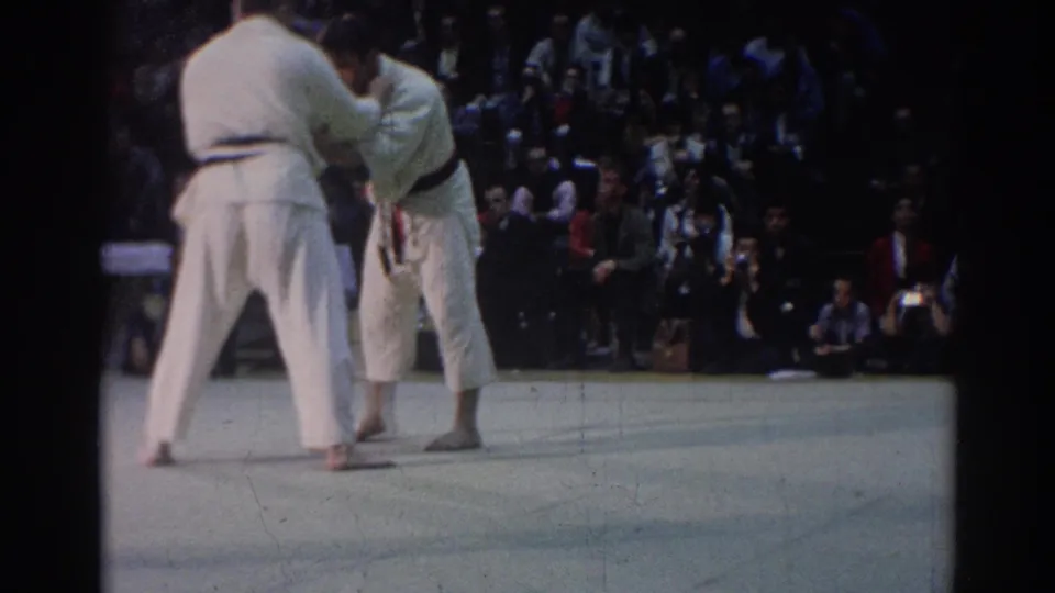 The match returns to an upright grip, showing two barefoot competitors in white uniforms before spectators in 1967 Los Angeles footage.