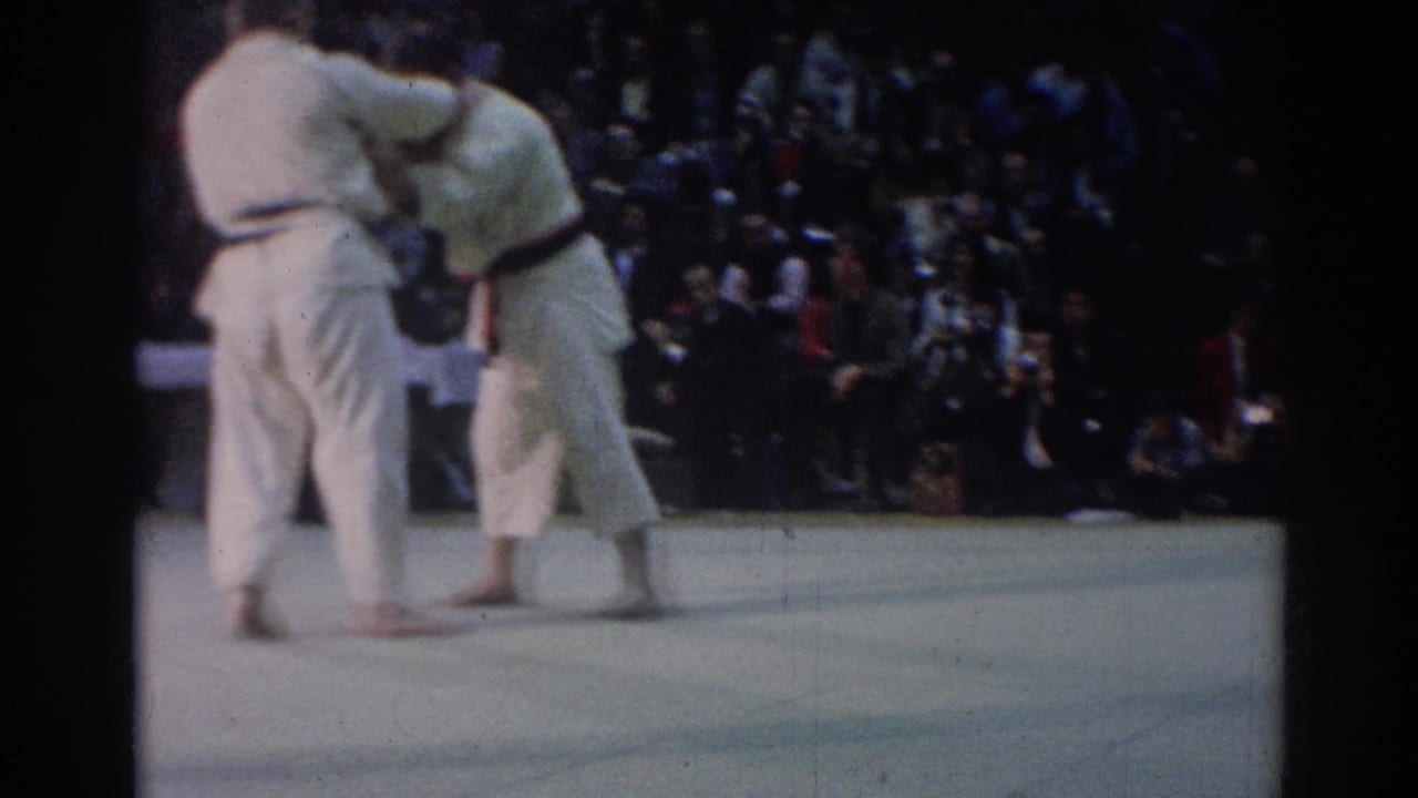 Two judo competitors in white gi grapple on a light mat before a seated crowd.