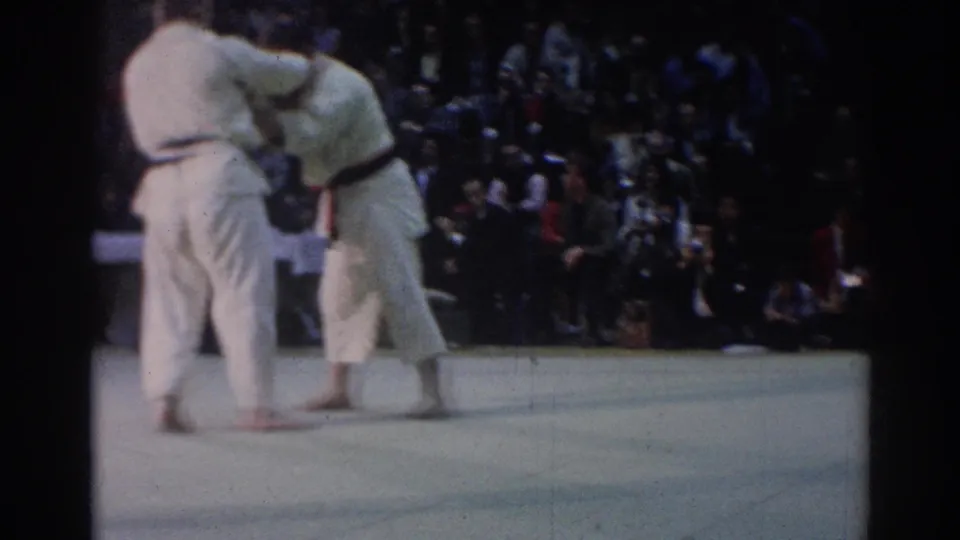 1967 Los Angeles judo competitors appear to grip each other on a light competition floor while spectators watch from the background.