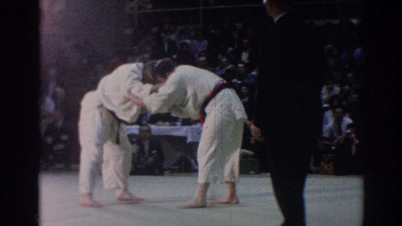The match continues as two judo competitors bend into a grip exchange on the mat, with a dark-clad official and spectators visible nearby.