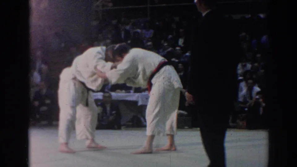 The match continues as two judo competitors bend into a grip exchange on the mat, with a dark-clad official and spectators visible nearby.