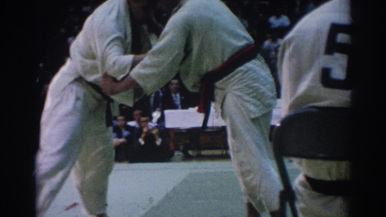 Two judo competitors in white gi grapple on a mat beside a seated official.