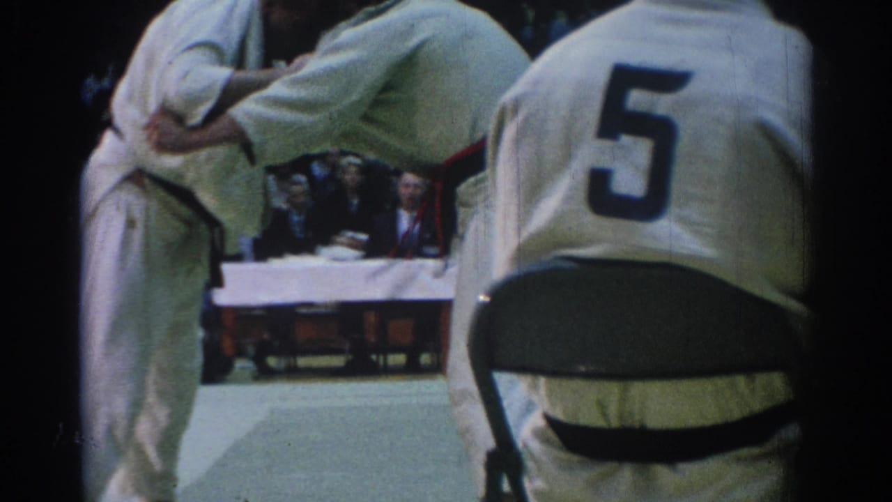 Two black-belt judo competitors continue grappling on an indoor mat in Los Angeles, with spectators and judges visible behind them.