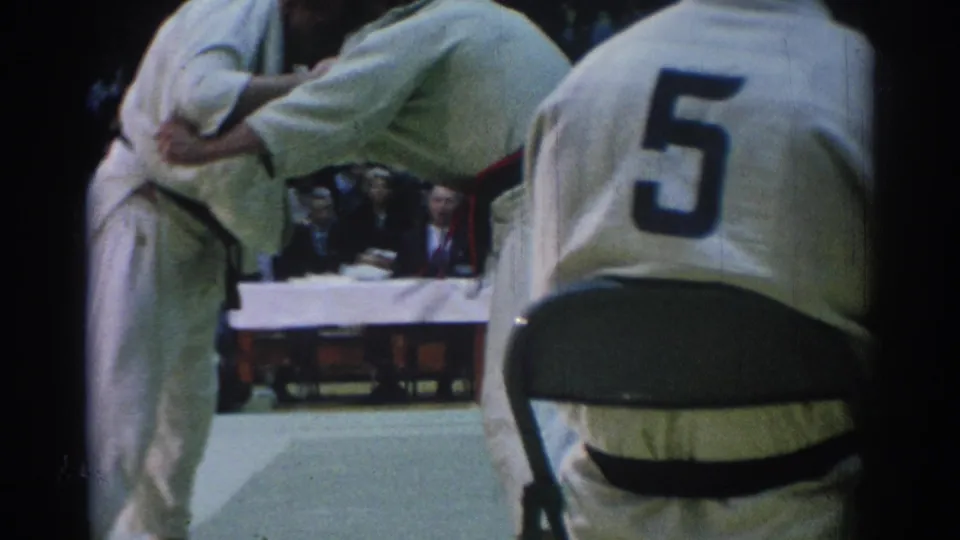 Two black-belt judo competitors continue grappling on an indoor mat in Los Angeles, with spectators and judges visible behind them.
