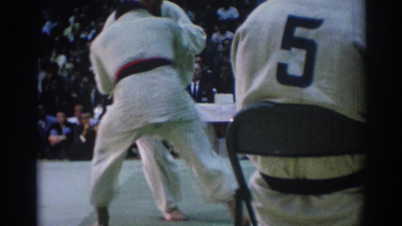 A competitor in a white gi pivots during a judo exchange as the foreground official and background spectators frame the 1967 match.