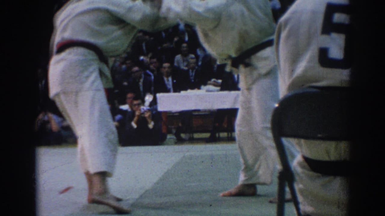 Near the end of the clip, the judo competitors lift and counterbalance each other on the mat with judges and spectators in the background.
