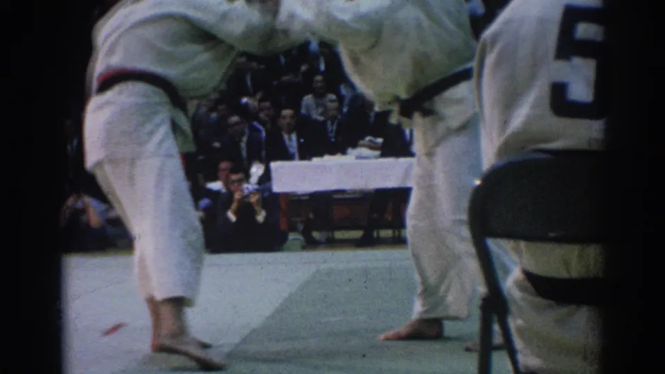 Near the end of the clip, the judo competitors lift and counterbalance each other on the mat with judges and spectators in the background.