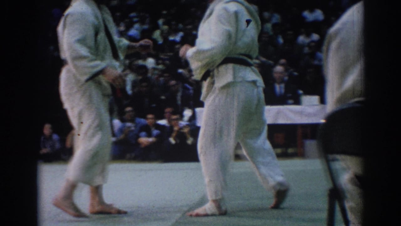 Two judokas in white gi stand on a mat before a seated audience.