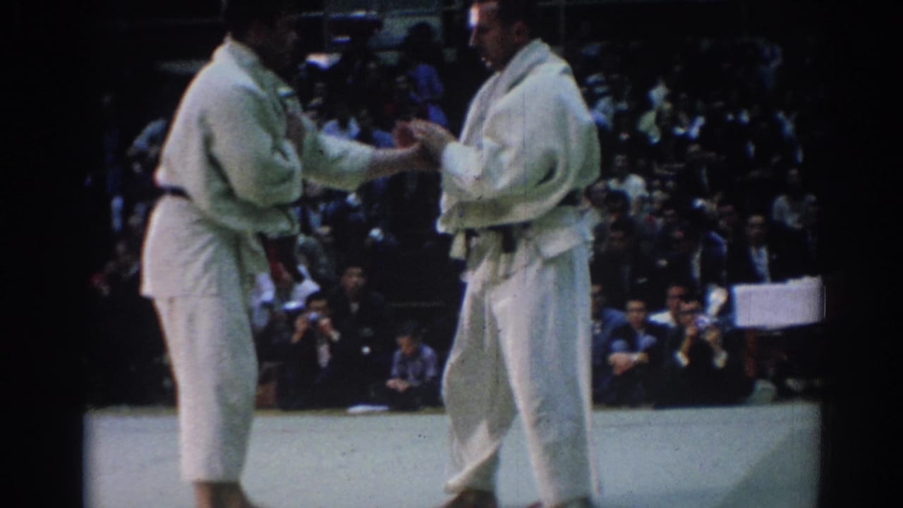 Two male judokas in white uniforms meet at the start of a match, with spectators and officials visible in the background.