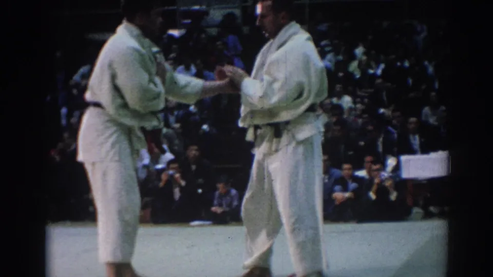 Two male judokas in white uniforms meet at the start of a match, with spectators and officials visible in the background.