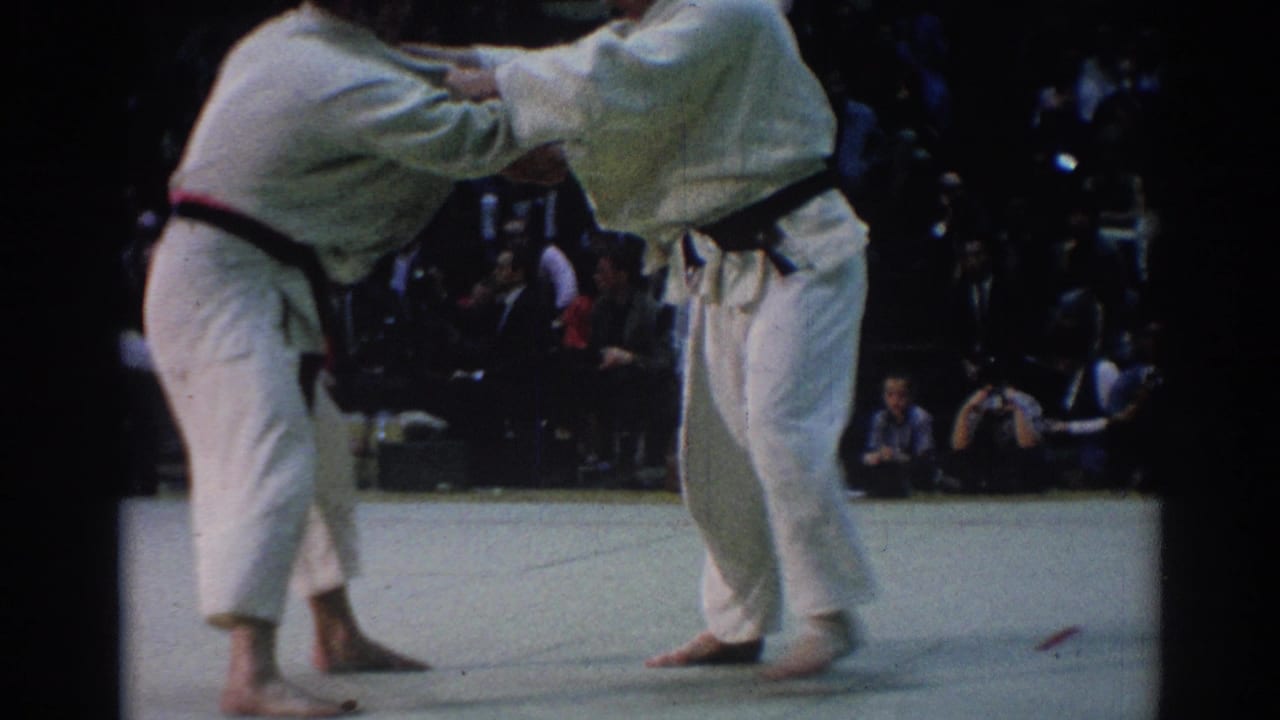 Two male judokas in white gi and black belts lean into a standing exchange as spectators watch from behind the mat.