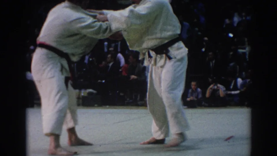 Two male judokas in white gi and black belts lean into a standing exchange as spectators watch from behind the mat.