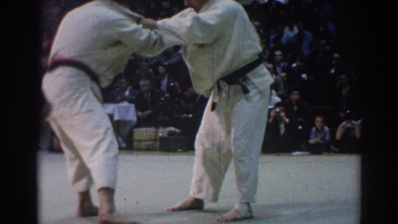 Two judo athletes in white gi grapple on an indoor mat before seated spectators.