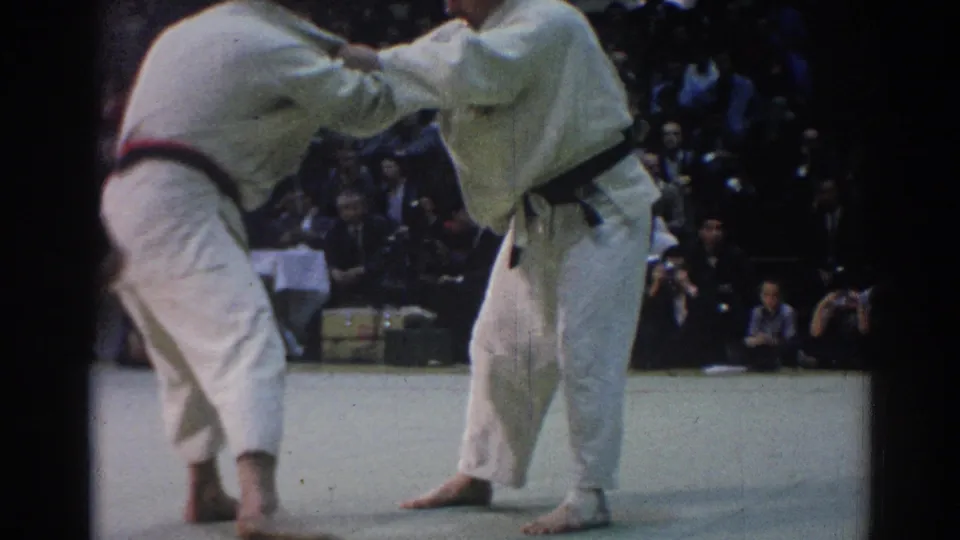 1967 Los Angeles 8mm frame of two judo competitors in white gi and black belts gripping each other on a competition mat.