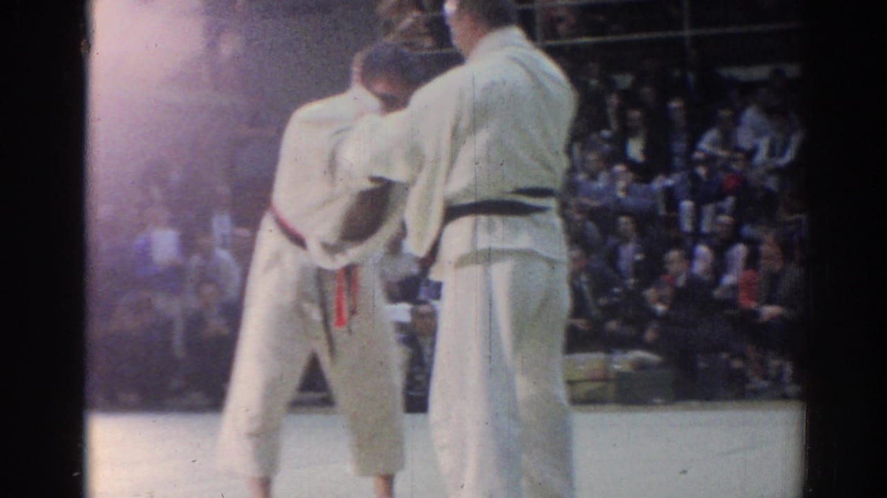 Two judo athletes in white uniforms appear between grips during a 1967 Los Angeles indoor competition, with spectators in the background.