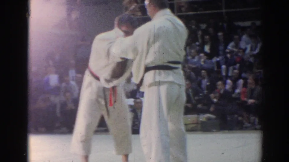 Two judo athletes in white uniforms appear between grips during a 1967 Los Angeles indoor competition, with spectators in the background.