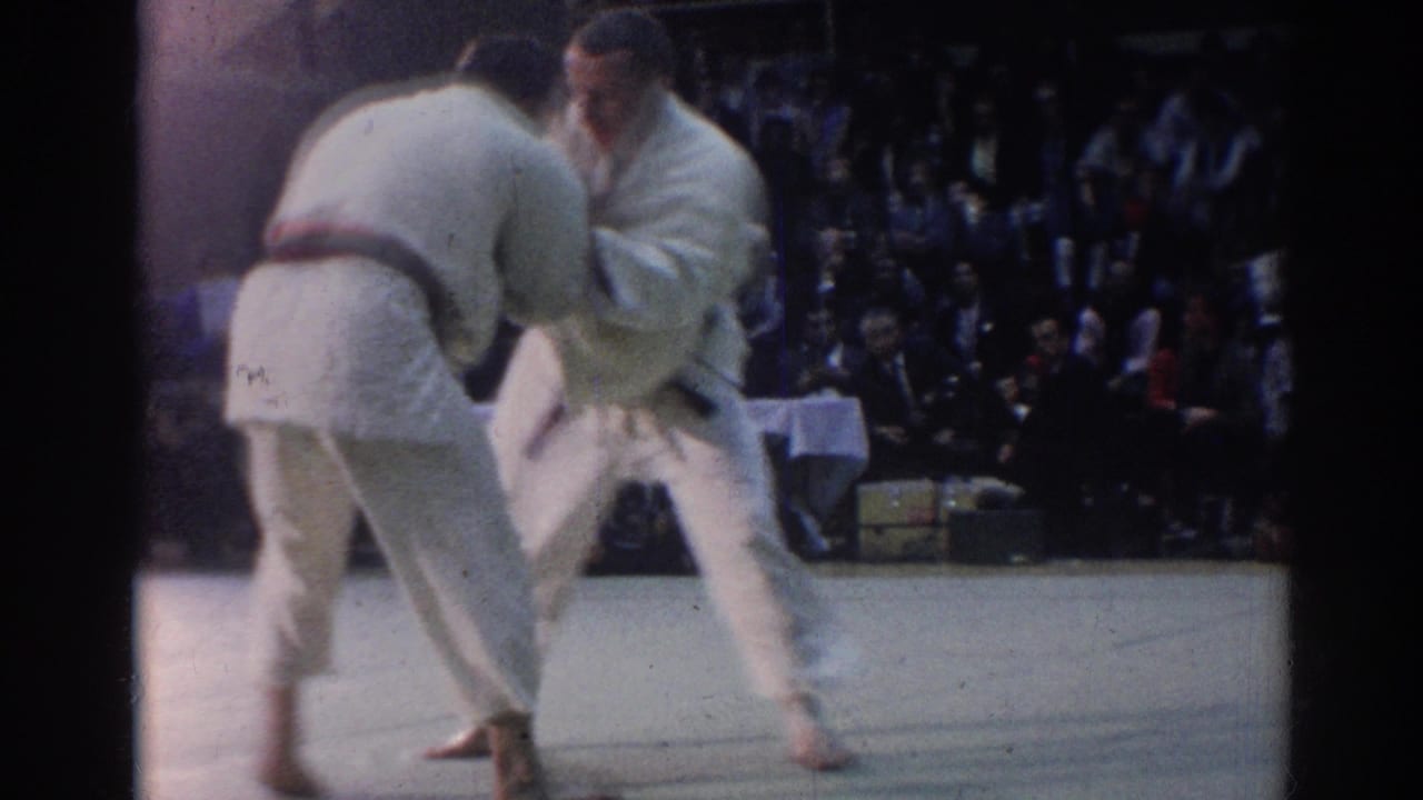 A 1967 Los Angeles judo match continues as two athletes lean forward in a close grip near seated observers and a table.