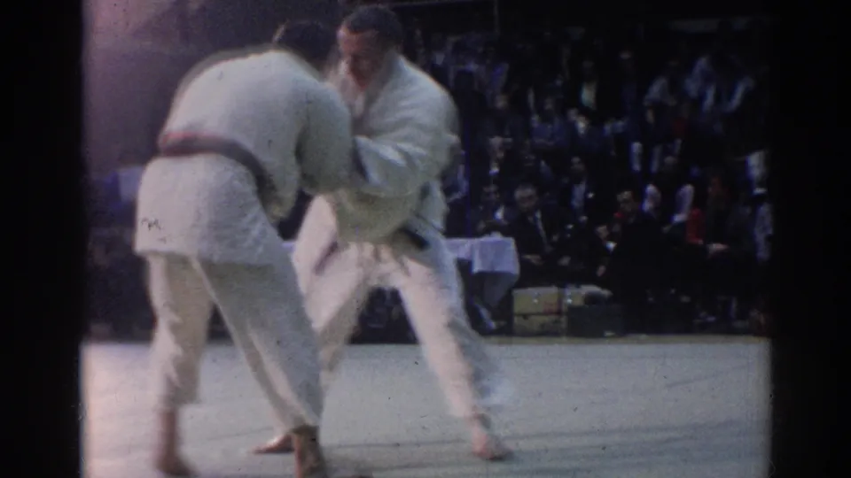 A 1967 Los Angeles judo match continues as two athletes lean forward in a close grip near seated observers and a table.
