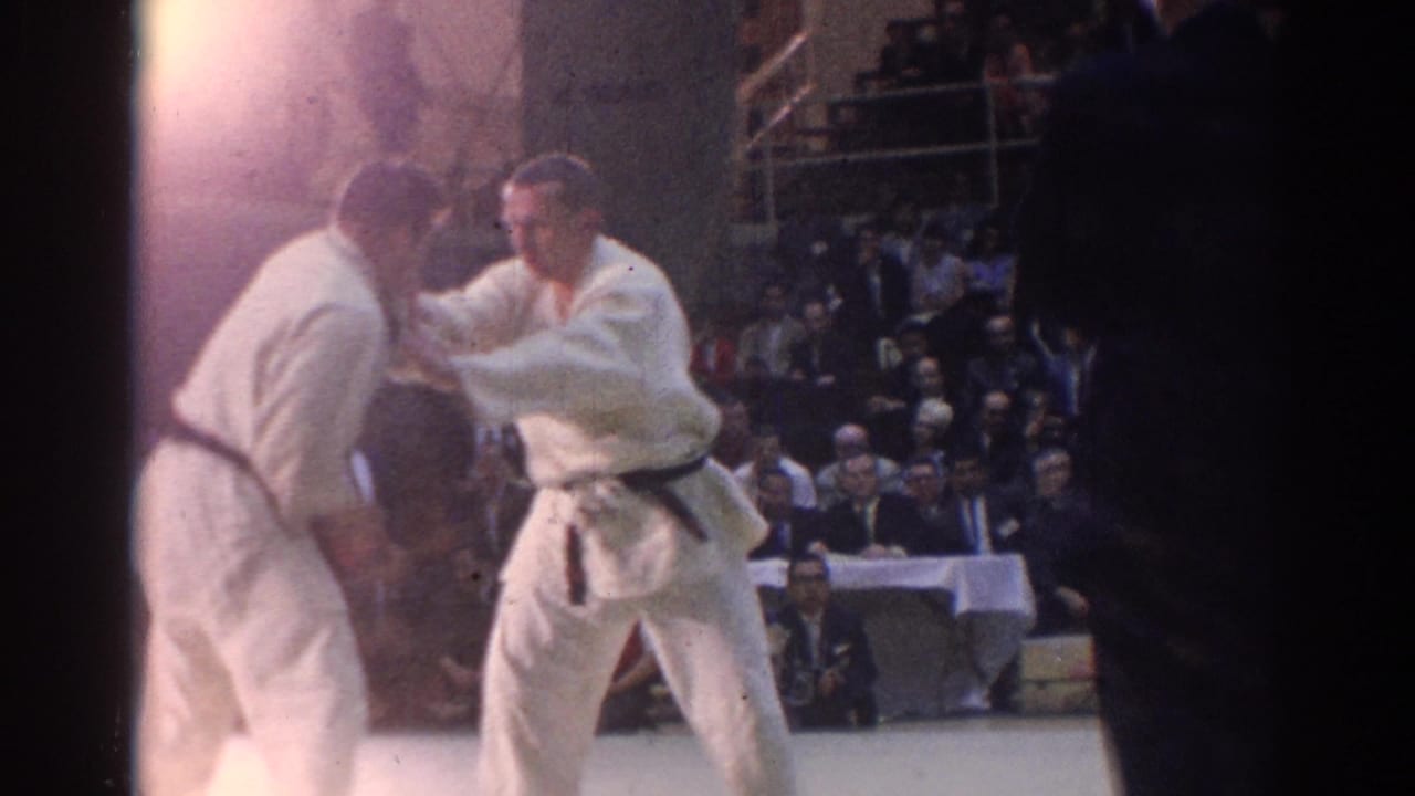 Two men in white gi grapple during an indoor karate match as spectators watch behind them.