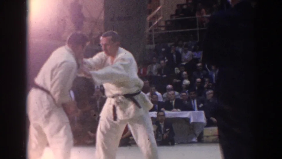 1967 Los Angeles 8mm frame of two male karate competitors in white gi on a mat, with a seated audience and judges' table in the background.