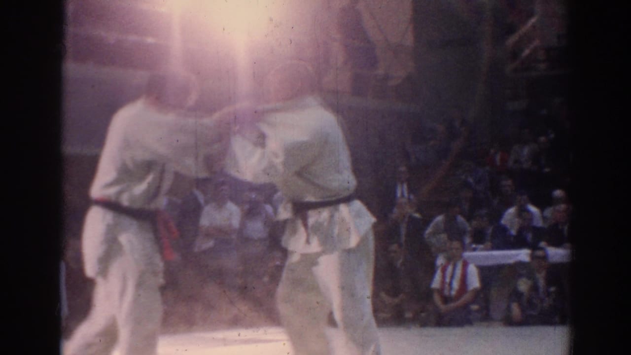 Soft 8mm frame from the Los Angeles karate match showing two adult competitors in white gi, partially obscured by flare and film exposure.