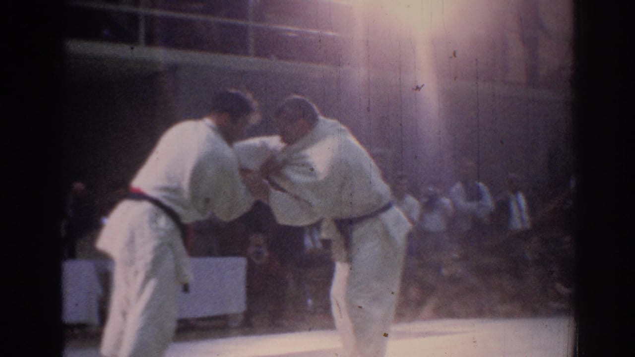 1967 Los Angeles indoor karate match frame with two adult competitors grappling, seen through soft focus, grain, and strong overhead glare.