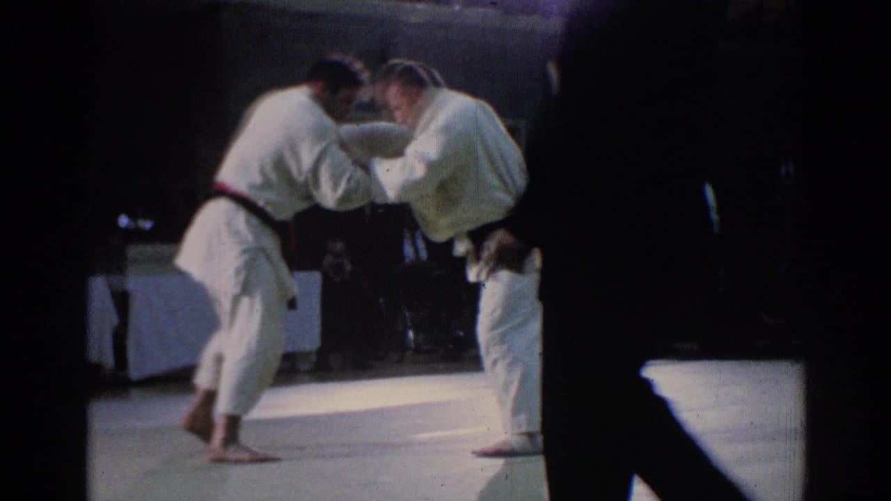 Late-1960s Los Angeles 8mm frame showing a referee observing two adult karate competitors in white gi during an indoor match.