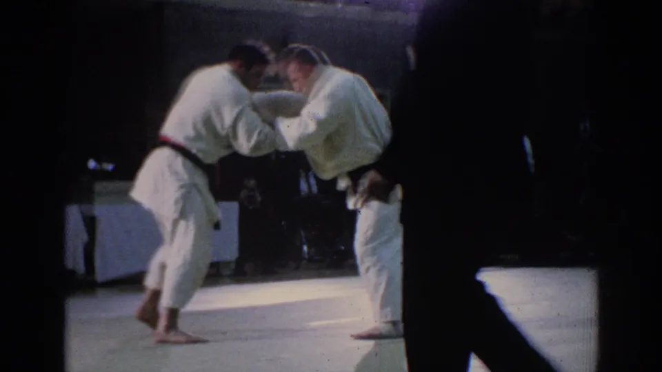 Late-1960s Los Angeles 8mm frame showing a referee observing two adult karate competitors in white gi during an indoor match.