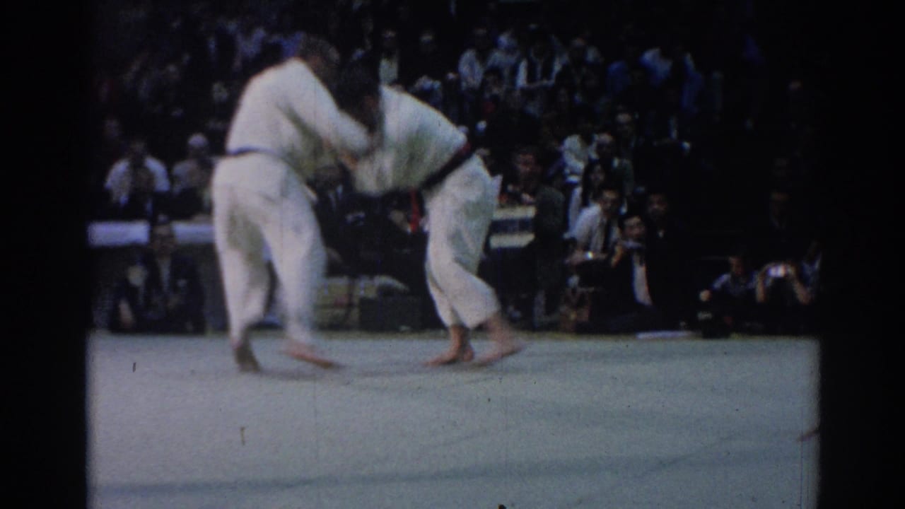 Two judo competitors in white uniforms grapple on a pale mat before seated spectators.