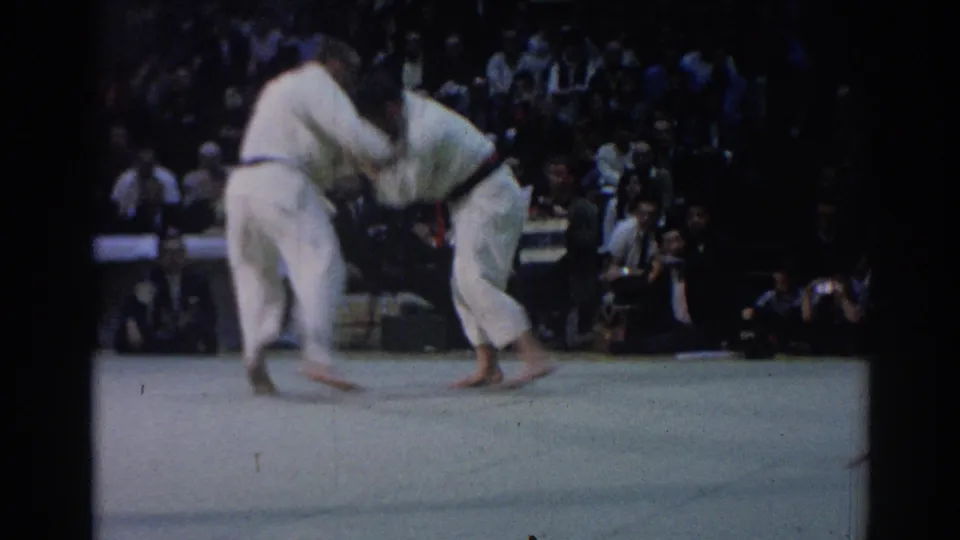 1967 Los Angeles 8mm frame of two judoka gripping each other on an indoor mat with spectators in the dim background.