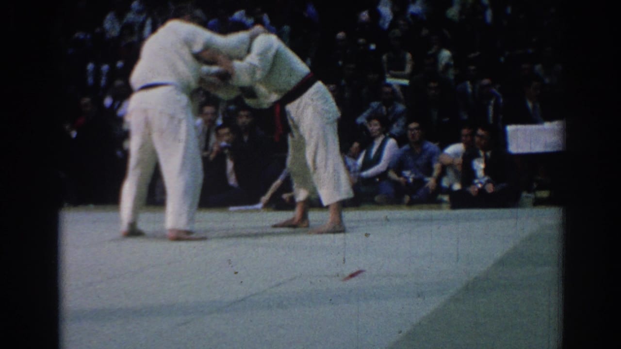 1967 Los Angeles 8mm frame of a judo exchange, with one competitor bent forward and the other holding position before spectators.