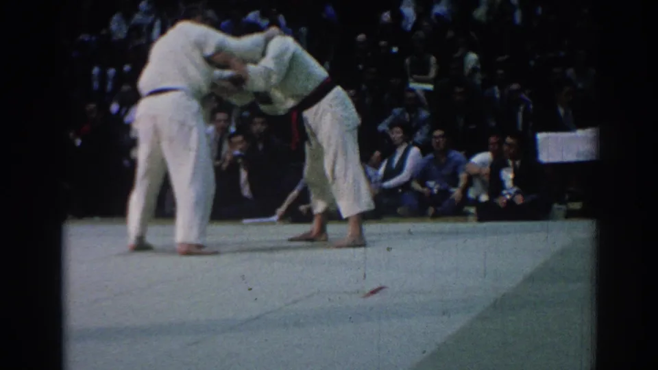 1967 Los Angeles 8mm frame of a judo exchange, with one competitor bent forward and the other holding position before spectators.