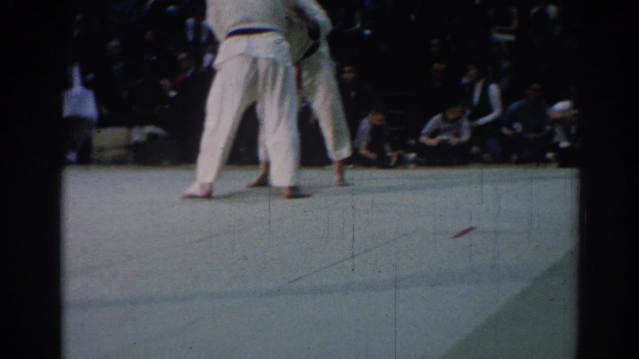 1967 Los Angeles 8mm frame showing the judo match continuing on a pale indoor mat with spectators along the far side.