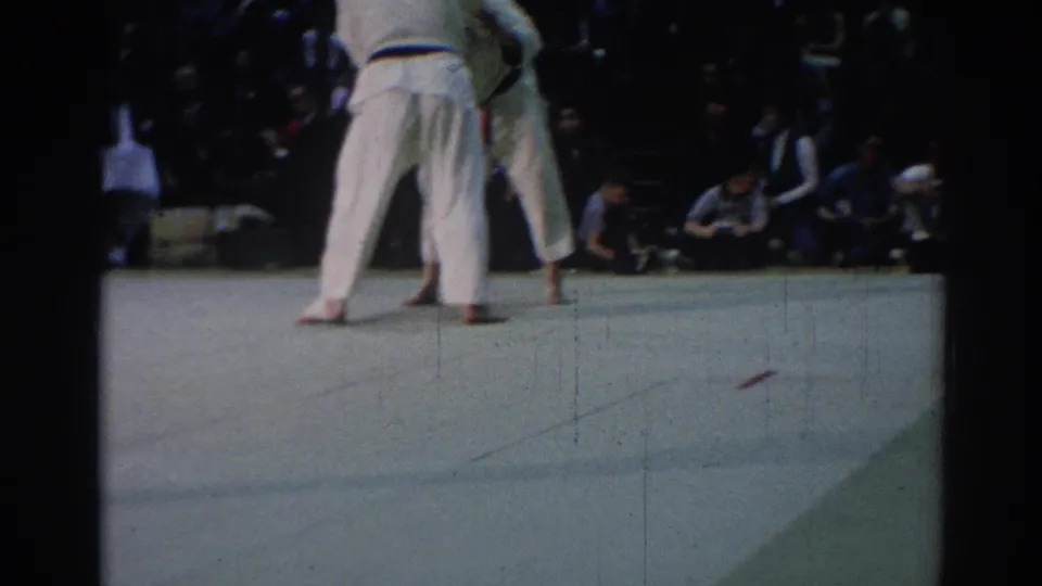 1967 Los Angeles 8mm frame showing the judo match continuing on a pale indoor mat with spectators along the far side.
