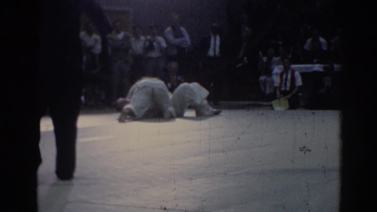 Two judo athletes grapple low on a mat while spectators sit in the background.