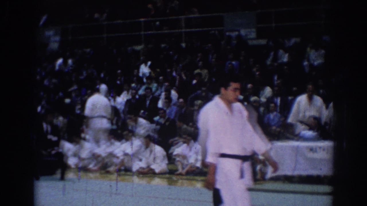 1967 Los Angeles judo competition scene with a standing competitor, seated athletes in white gi, and a crowded arena background.