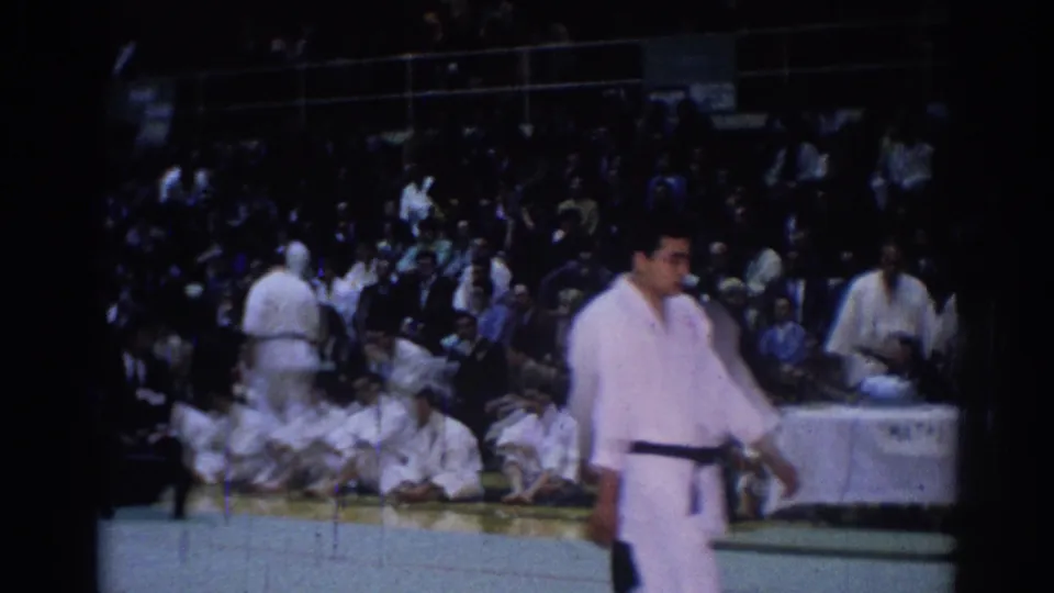 1967 Los Angeles judo competition scene with a standing competitor, seated athletes in white gi, and a crowded arena background.