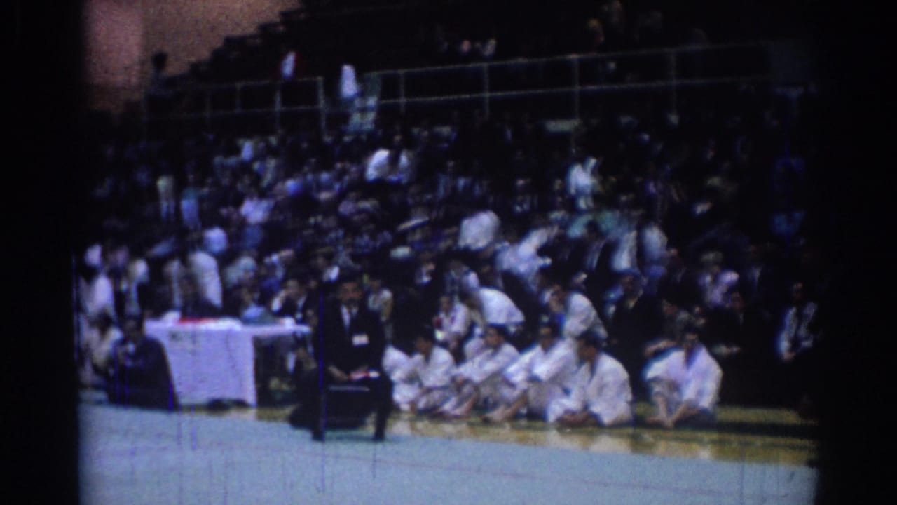 1967 Los Angeles judo competition view showing a dark-suited official beside seated athletes and a large audience in the stands.